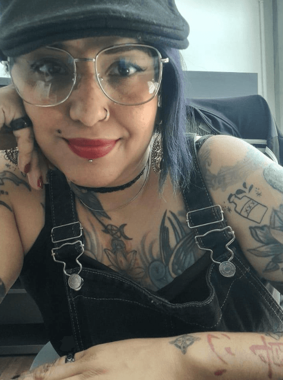 Plano medio de Alejandra Collado, tallerista de Ciempiés. Alendra tiene cabello oscuro con reflejos violáceos, usa gafas, tiene un argolla en la nariz y lleva puesta una gorra. Mira directamente a cámara y sonríe. Luce infinidad de tatuajes.