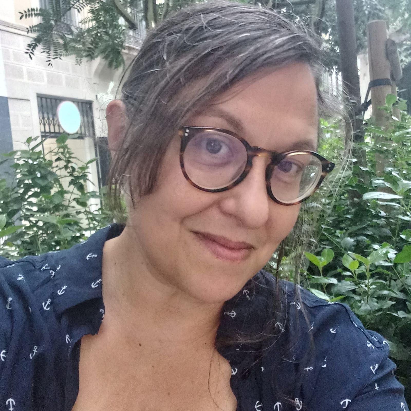 Primer plano de medio perfil de Natalia Renzi. Natalia tiene el cabello lacio de color castaño, en la foto lo lleva recogido con un mechón suelto que enmarca el lado derecho de su rostro. Usa gafas y lleva una camisa azul a lunares blancos.