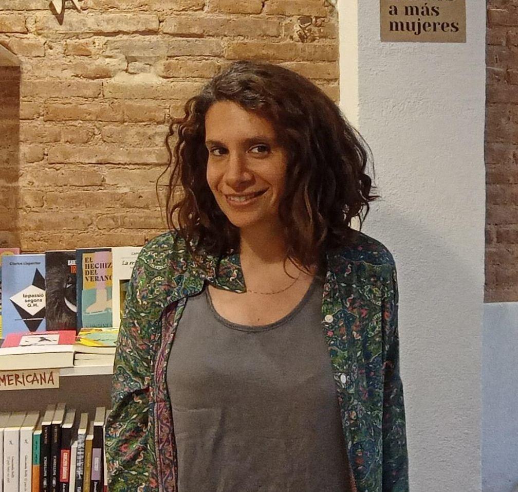 Plano medio de Paola Bolelli, tallerista de Ciempiés. Paola es una mujer de cabellos oscuros y rizados, que le llegan hasta los hombros. En la pared detrás de ella se observan varios libros. Viste una blusa en tonos verdes que lleva abierta y deja ver una camiseta gris.