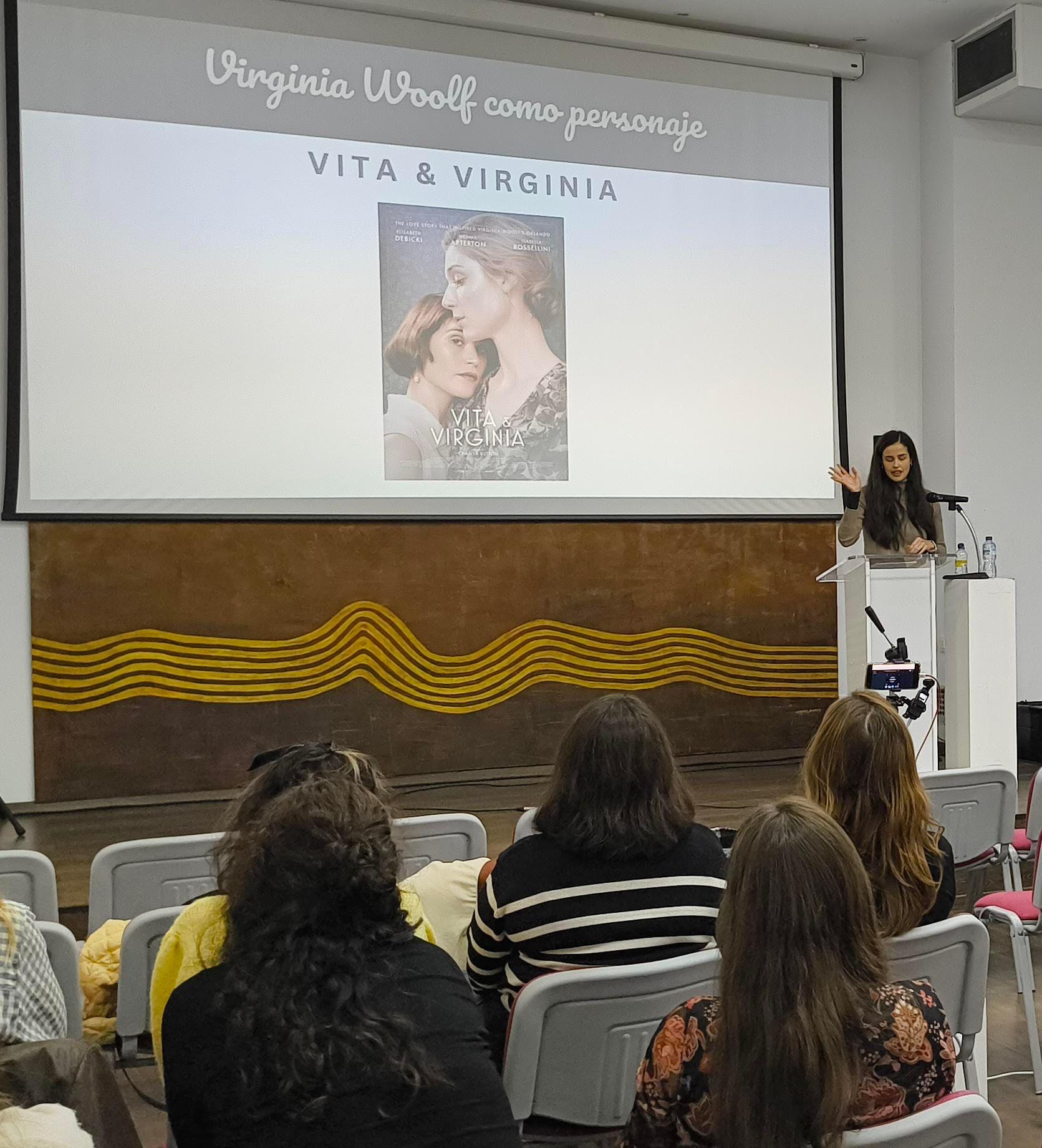 Una fotografía que muestra a Sabina Torres de pie frente a un atril con micrófono y con una pantalla a sus espaldas. Es un plano desde lejos y se ve parte del público.