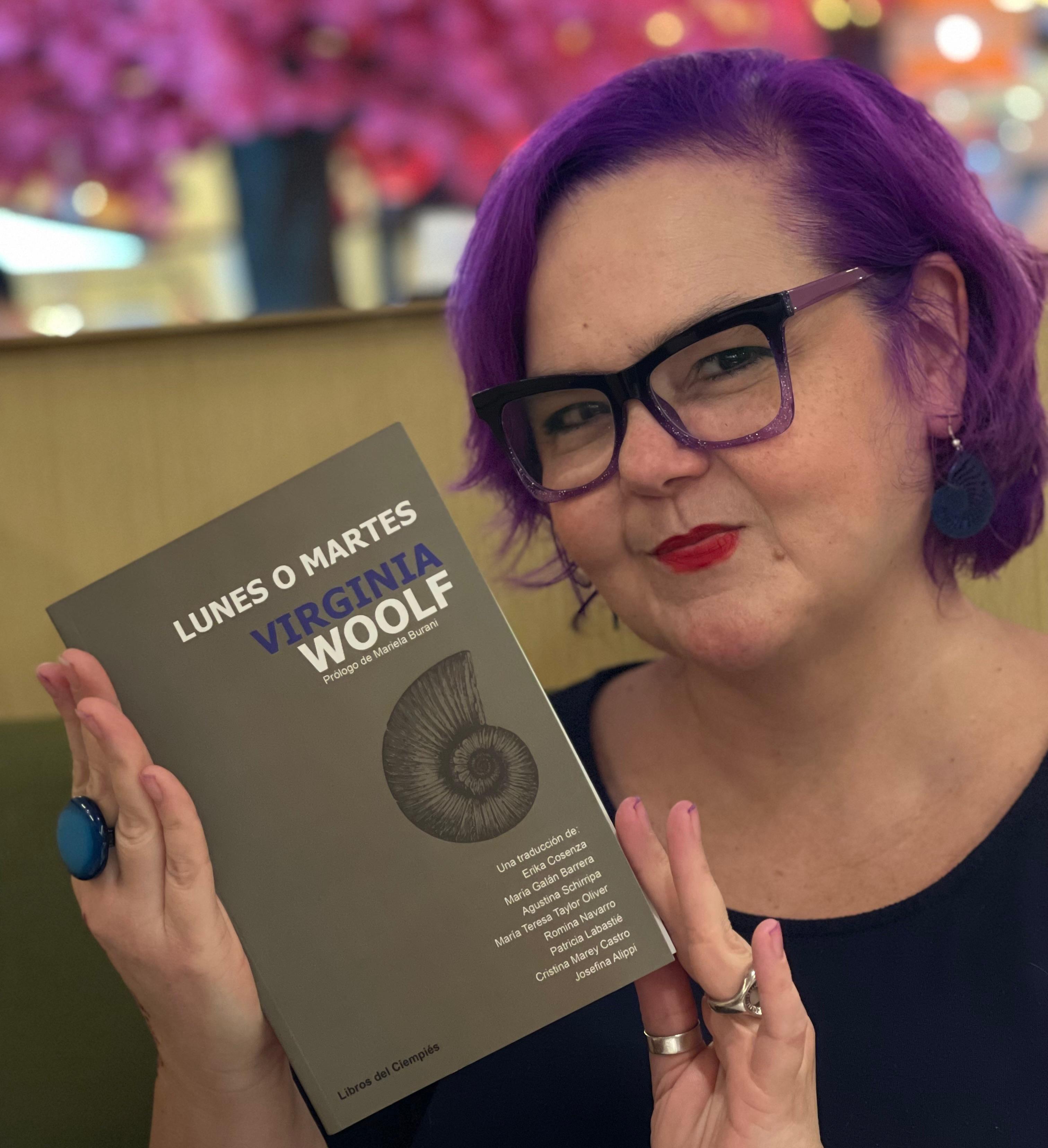 Plano medio de María Teresa Taylor Oliver. Tiene el cabello corto y de color violeta. Lleva gafas de marco grueso y negro. En la foto sostiene un ejemplar del libro "Lunes o martes", que publicó Libros del Ciempiés con María Teresa como una de las traductoras.