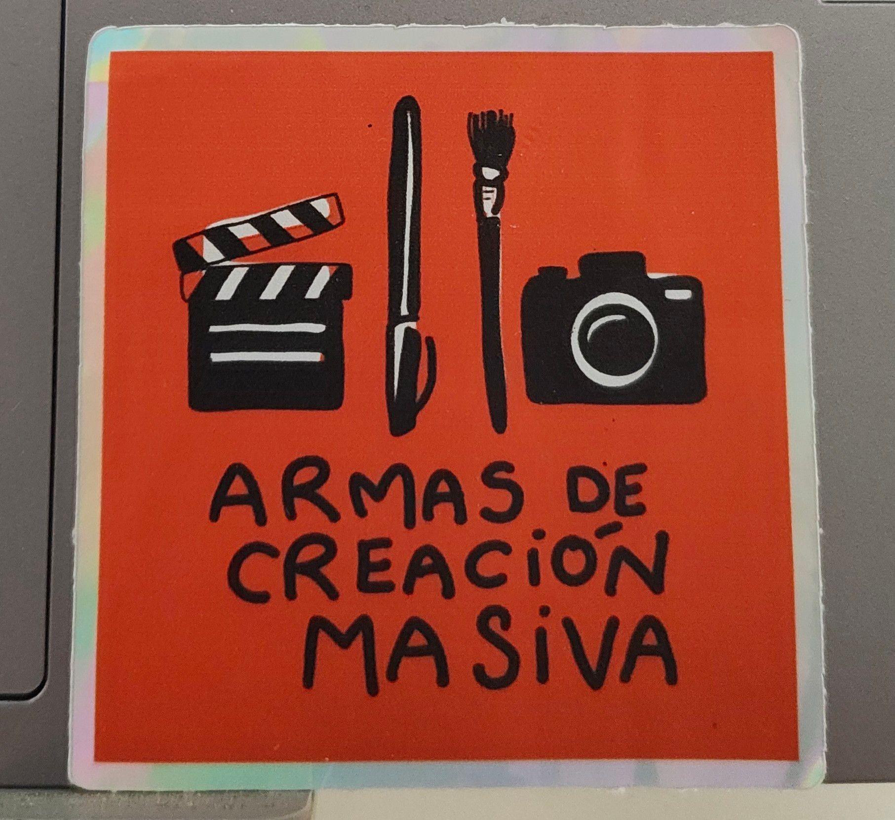 Fotografía de una pegatina que, sobre fondo rojo, muestra las ilustraciones en negro de una claqueta de cine, un bolígrafo, un pincel y una cámara fotográfica. Debajo el texto pone «armas de creación masiva».