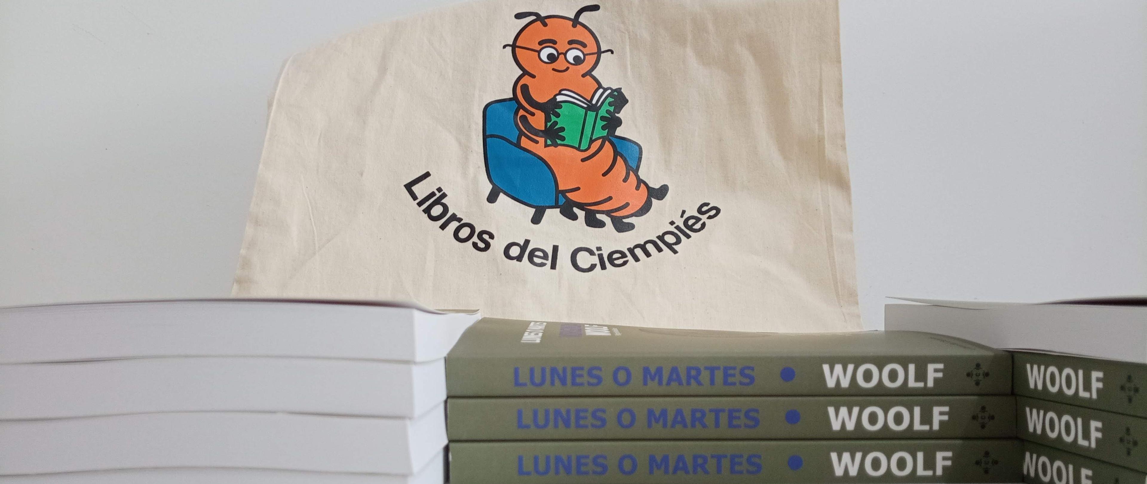 Una tote bag color crudo con la imagen del »Ciempiés lector» debajo de la ilustración dice «Libros del Ciempiés». La bolsa está por detrás de una pila de ejemplares del libro «Lunes o martes», de Viriginia Woolf, editado por Libros del Cimpiés para su colección «Clásicos Accesibles».