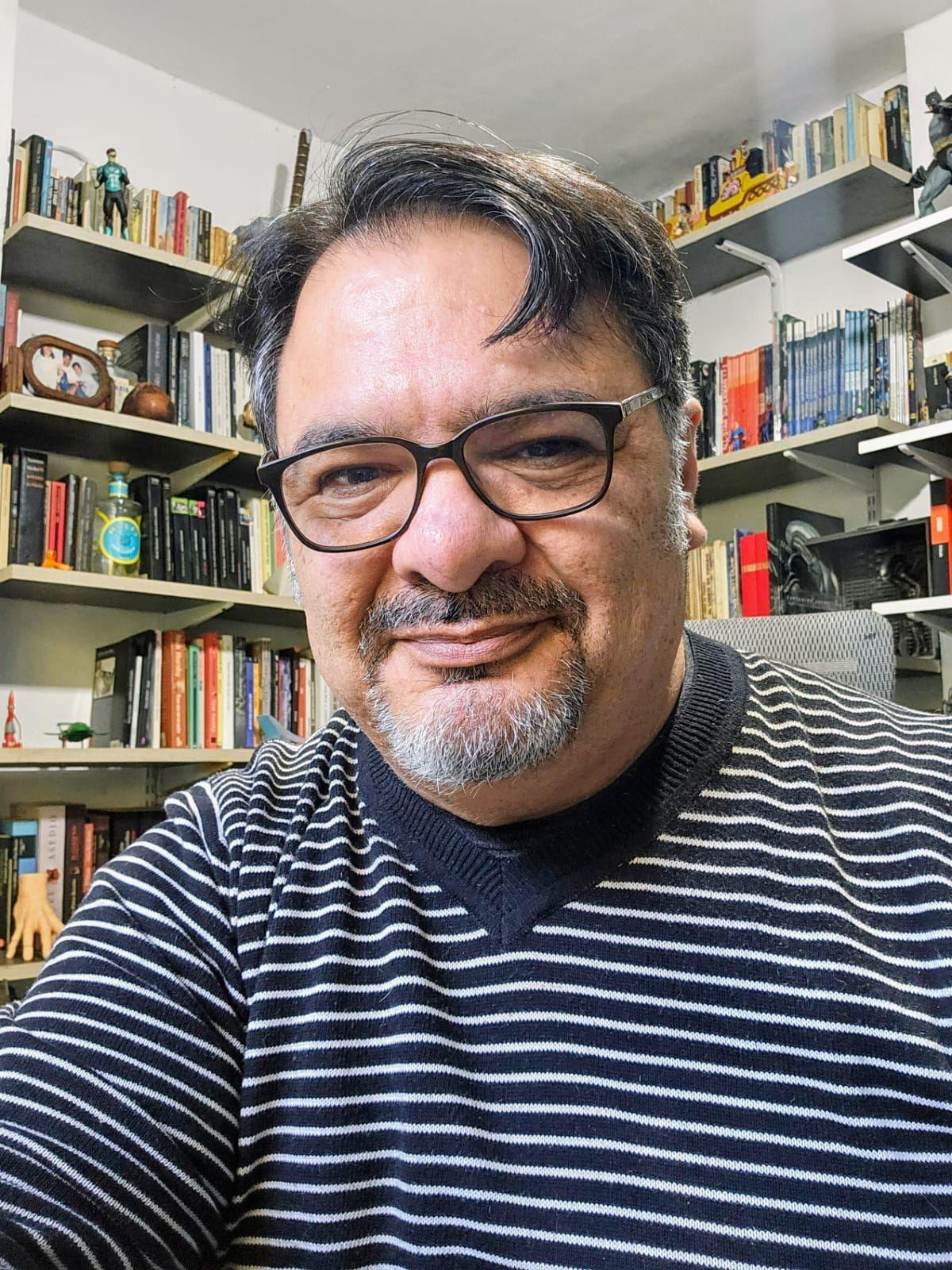 Plano medio de Marcelo Cabrera. Tiene el cabello corto, de color castaño, y barba entrecana. Lleva gafas. Detrás de él, puede verse una estantería repleta libros, recuerdos y adornos que llega hasta el techo.